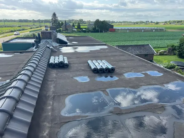 Vóór het werk: groot plat dak met plassen water en rollen dakbedekking klaarliggend