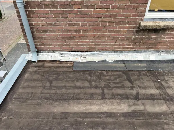 Vóór het werk: verweerde bruine dakbedekking met opstand langs de gevel en regenpijp